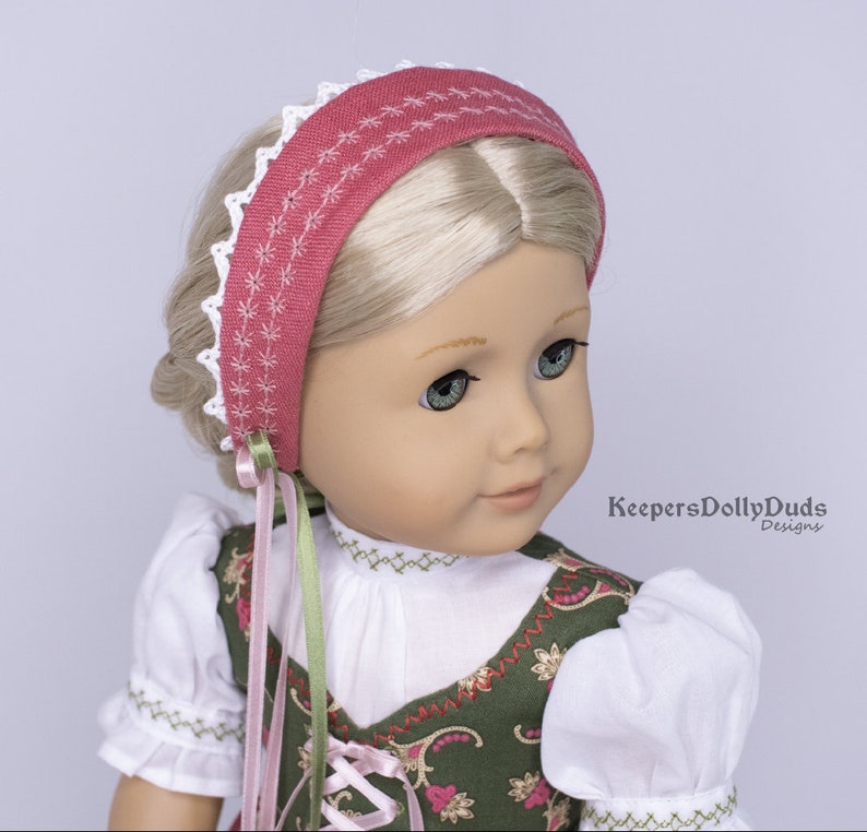 May include: A doll wearing a white blouse with green and gold embroidery, a pink and green ribbon, and a pink headband with white lace trim. The headband has a white embroidered design. The doll has blonde hair and blue eyes. The text "KeepersDollyDuds Designs" is visible in the lower right corner of the image.