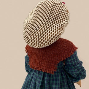 May include: A doll wearing a white mesh hairnet with a burgundy bow, a brown shawl, and a green and blue plaid dress. The doll's name, "Flossie Potter", is written on the dress.