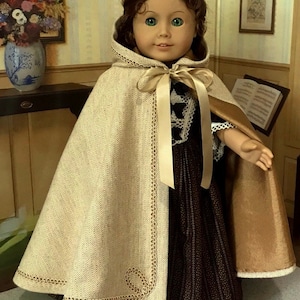 May include: A doll wearing a brown and white dress with a beige cape and a white lace bonnet. The cape has gold trim and a satin ribbon tied at the neck. The doll is standing in front of a wooden table with a vase of flowers on it.