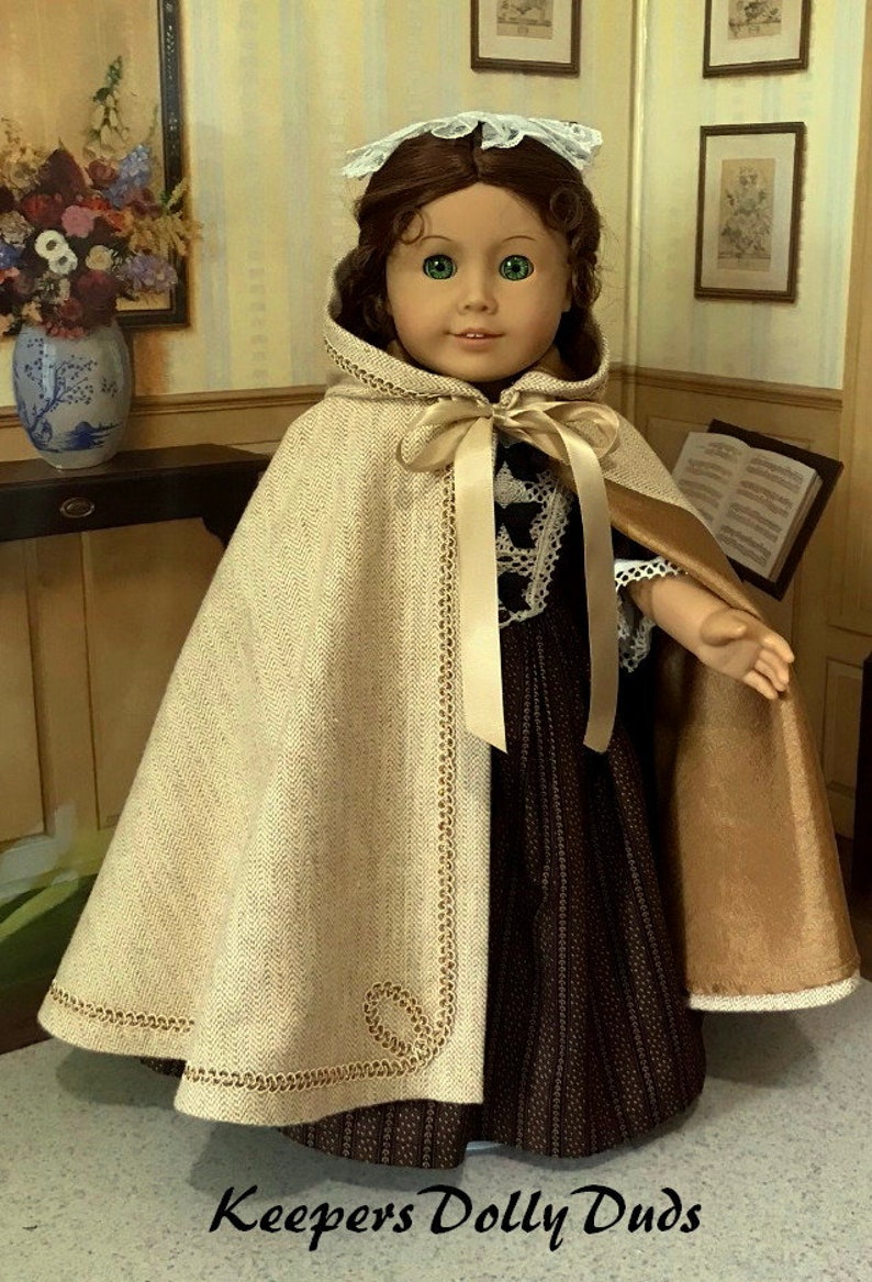 May include: A doll wearing a brown and white dress with a beige cape and a white lace bonnet. The cape has gold trim and a satin ribbon tied at the neck. The doll is standing in front of a wooden table with a vase of flowers on it.