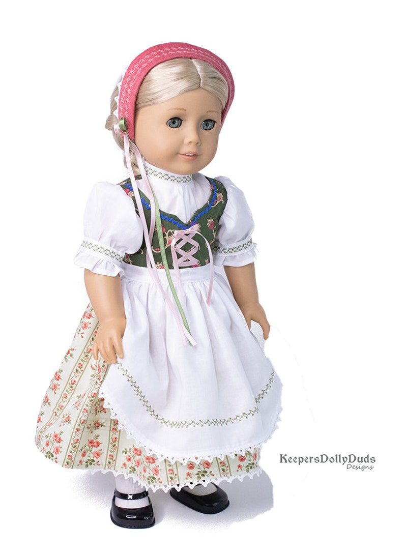 May include: A doll wearing a green and white dirndl with a pink ribbon and a pink headband. The doll has blonde hair and blue eyes. The doll is wearing black shoes. The doll is standing on a white background. KeepersDollyDuds Designs.