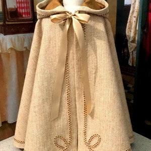 May include: A beige hooded cape with a gold satin ribbon and gold trim. The cape is made of a textured fabric and has a fitted bodice. The cape is on a mannequin and the text "Keepers Dolly Duds" is at the bottom of the image.