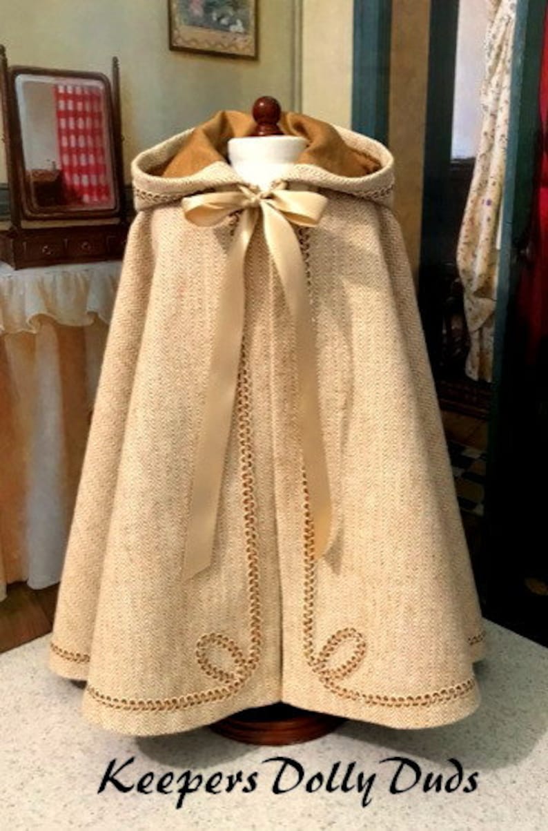 May include: A beige hooded cape with a gold satin ribbon and gold trim. The cape is made of a textured fabric and has a fitted bodice. The cape is on a mannequin and the text "Keepers Dolly Duds" is at the bottom of the image.