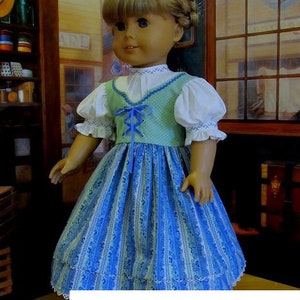 May include: A doll wearing a blue and green dress with a white blouse and a blue ribbon. The dress has a lace trim and a striped pattern. The doll is standing in front of a wooden shelf with various items on it.