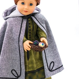 May include: A doll wearing a gray hooded cape with black trim, a green dress with a black lace trim, and black shoes. The doll is holding a small bird's nest with colorful eggs and berries. KeepersDollyDuds Designs &copy;2021 Eve Coleman. All Rights Reserved.