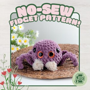 May include: A purple crocheted spider fidget toy with orange eyes and fluffy white accents. The toy is on a woven mat, with the text "NO-SEW FIDGET PATTERN!" above it. A PDF download icon is in the lower right corner.
