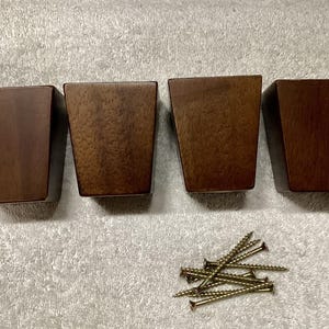 May include: Four dark brown wooden furniture legs with a tapered design. The legs are square at the top and wider at the base. A small pile of gold-colored screws is also included.