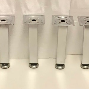 May include: Four silver metal furniture legs with a square base and a round foot. The legs are approximately 6 inches tall.