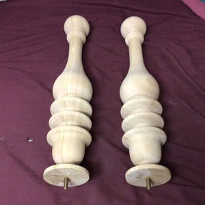 May include: Two wooden finials with a bulbous shape and a series of rings. The finials are light brown and have a threaded metal rod at the bottom.