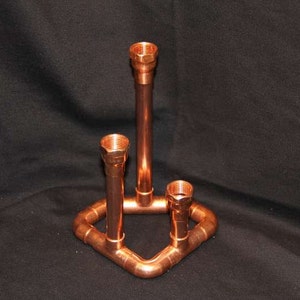 Puede incluir: Un portavelas de tubería de cobre con tres portavelas. El portavelas está hecho de tubería de cobre y tiene un aspecto rústico.