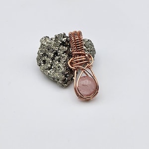 May include: A handcrafted pendant featuring a teardrop-shaped, light pink gemstone encased in copper wire. The wire is intricately woven, creating a cage-like design. The pendant rests on a textured, gray mineral, set against a white background.