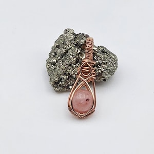 May include: A handcrafted pendant featuring a pink gemstone encased in copper wire. The wire is intricately woven around the stone, creating a decorative cage. The pendant is displayed against a textured, gray stone.