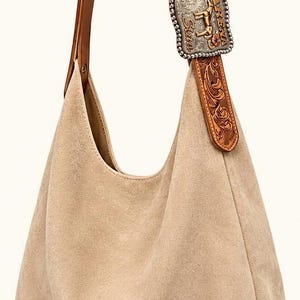 May include: A beige suede hobo bag with a brown leather strap. The strap is connected to a decorative silver buckle with the text "Z Farina Arena" and floral and horse details. The bag has a soft, slouchy design.