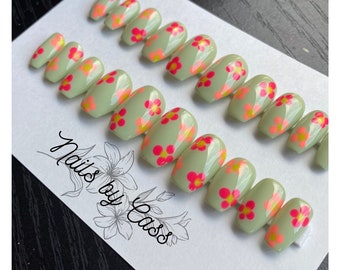 May include: A set of oval-shaped press-on nails in a light green hue, adorned with vibrant pink and yellow floral designs. The nails are arranged on a white card with the text "Nails by Cass" in cursive.