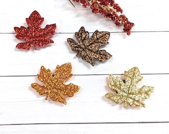 May include: Four maple leaf-shaped ornaments covered in glitter, featuring one red, one dark brown, one orange, and one gold leaf. Each leaf has a detailed vein pattern and a textured, sparkling surface, arranged with a sprig of red berries.