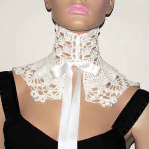 Puede incluir: Collar de encaje blanco de ganchillo con una cinta de sat&eacute;n blanca atada en un lazo. El collar tiene detalles de perlas.