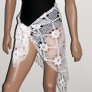 May include: A white, open-weave crochet sarong with a floral pattern. The sarong is tied at the waist and drapes over the legs. The crochet work is intricate, with a delicate, lacy appearance. The sarong is worn over a black swimsuit.