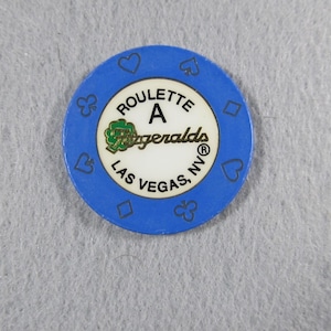May include: A blue casino roulette chip with a white center. The center has the text "ROULETTE A Fitzgeralds LAS VEGAS, NV" in black and gold. The outer ring has suit symbols.