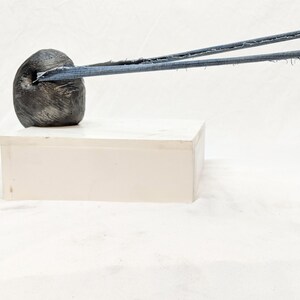 May include: A gray, abstract sculpture with a rough, textured surface is connected to a round, gray object by a blue cord. The sculpture is on a white surface.
