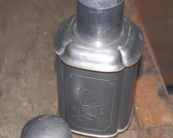 May include: A pewter tea caddy with a rectangular body and a cylindrical neck. The caddy features a decorative floral design on the front panel and a separate, rounded lid. The metal has a dark, matte finish.
