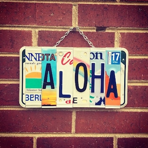 May include: A sign spelling "ALOHA" made from repurposed license plates. The plates are from different states and have various designs and colors. The sign is hanging on a brick wall.