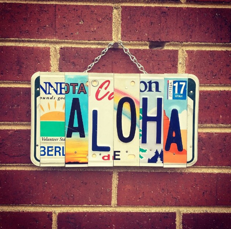 May include: A sign spelling "ALOHA" made from repurposed license plates. The plates are from different states and have various designs and colors. The sign is hanging on a brick wall.