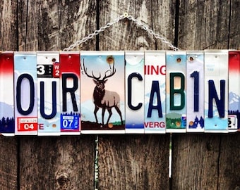 May include: A sign made of license plates that reads "OUR CABIN" with a picture of an elk in the middle. The sign is hanging on a wooden fence.
