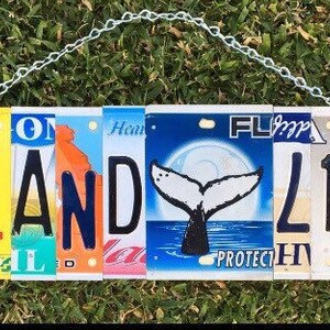 May include: A white sign with the words "Island Life" spelled out using colorful license plates. The sign is hanging from a chain and has a whale tail image on one of the plates.