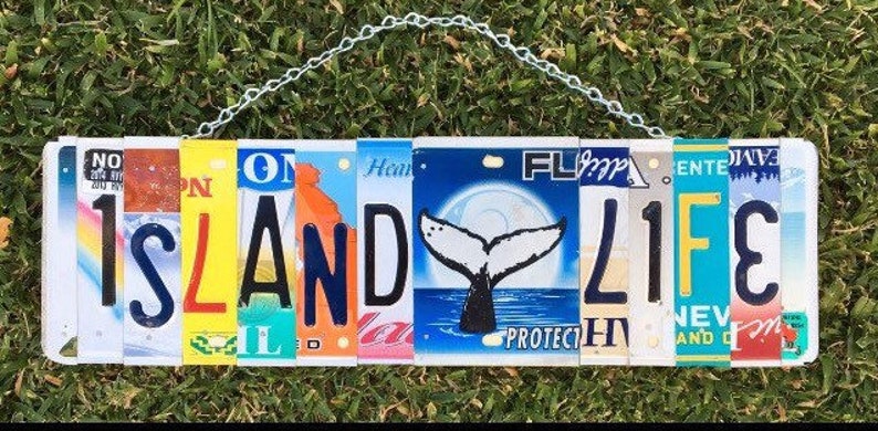 May include: A white sign with the words "Island Life" spelled out using colorful license plates. The sign is hanging from a chain and has a whale tail image on one of the plates.