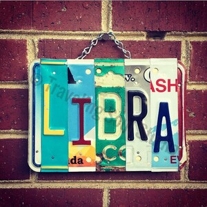May include: A sign spelling out the word "LIBRA" made from repurposed license plates. The letters are in different colors and styles, with some showing signs of wear and tear. The sign is hanging from a chain on a brick wall.