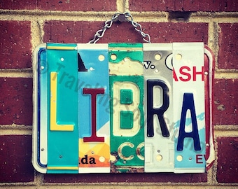 May include: A sign spelling out the word "LIBRA" made from repurposed license plates. The letters are in different colors and styles, with some showing signs of wear and tear. The sign is hanging from a chain on a brick wall.