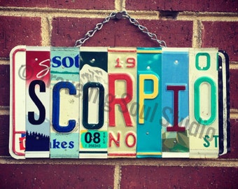 May include: A colorful sign spelling out "SCORPIO" made from repurposed license plates. The sign is hanging from a chain and is against a brick wall.