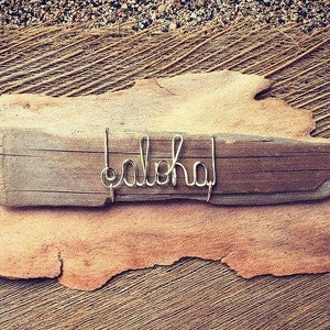 May include: A wooden sign with the word "aloha" in silver wire. The sign is placed on a piece of light brown bark, set against a background of textured, natural materials. The overall aesthetic suggests a beach or tropical theme.