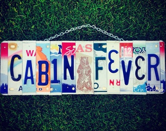 May include: A sign made of license plates spelling out "CABIN FEVER" with a bear image in the middle. The sign is hanging from a chain.