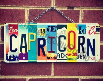 May include: A colorful sign spelling out "CAPRICORN" made from vintage license plates. The sign is hanging from a chain on a brick wall.