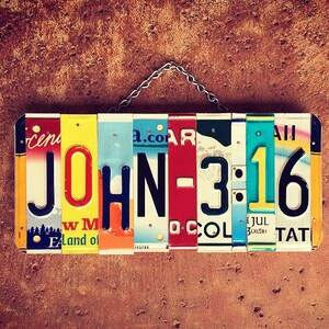 以下が含まれることがあります： さびた茶色の壁に、「JOHN-3:16」と綴られたナンバープレートでできたカラフルな看板が掛かっています。