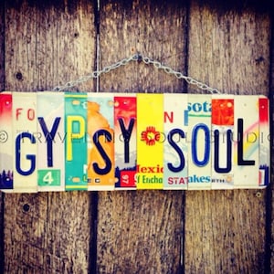 May include: A colorful sign made from license plates spelling out "GYPSY SOUL". The sign is hanging from a chain on a wooden background.