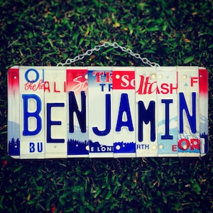 May include: A personalised name sign made from repurposed licence plates. The name "BENJAMIN" is spelled out in blue and black letters on a white background. The licence plates are arranged in a horizontal row and have red, white and blue colours.