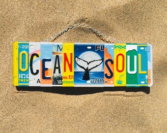 May include: A colorful sign spelling "OCEAN SOUL" made from repurposed license plates. The sign features a whale tail graphic and is hanging from a chain.