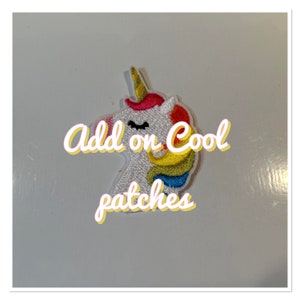 Peut inclure: Un patch thermocollant en forme de licorne blanche avec une crinière et une corne arc-en-ciel. Le patch dit "Add on Cool Patches" en texte jaune.