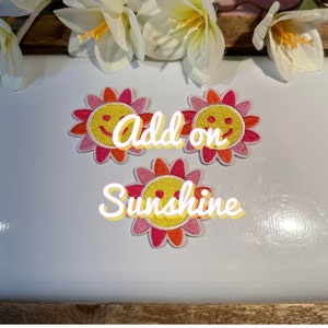 Puede incluir: Tres parches bordados para planchar en forma de sol amarillo sonriente con pétalos rosas y naranjas. El texto "Add on Sunshine" está escrito debajo de los parches.