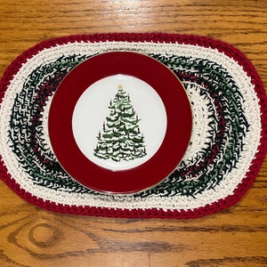 May include: A red and white crocheted oval placemat with a green and white design. A red and white plate with a gold rim and a Christmas tree design is on top of the placemat.