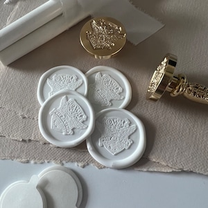 Könnte beinhalten: Vier weiße Wachssiegel mit einem geflügelten Emblem und dem Text "OWLC POST SERVICE" in einem kreisförmigen Design. Ein goldener Wachssiegel-Stempel mit einem geflügelten Emblem ist ebenfalls abgebildet.