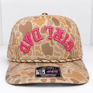 K&ouml;nnte beinhalten: Eine Baseballkappe mit Tarnmuster und den Worten "DAD GIRL" in leuchtendem Pink gestickt. Die Kappe hat einen gebogenen Schirm und eine Mesh-R&uuml;ckseite. Die Kappe hat ein Etikett mit der Aufschrift "EST. 2023 10 HATS WATERPROOF ADJUSTABLE/SURVEY".