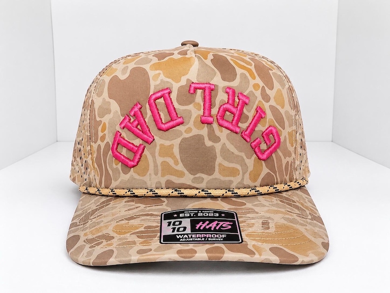 K&ouml;nnte beinhalten: Eine Baseballkappe mit Tarnmuster und den Worten "DAD GIRL" in leuchtendem Pink gestickt. Die Kappe hat einen gebogenen Schirm und eine Mesh-R&uuml;ckseite. Die Kappe hat ein Etikett mit der Aufschrift "EST. 2023 10 HATS WATERPROOF ADJUSTABLE/SURVEY".