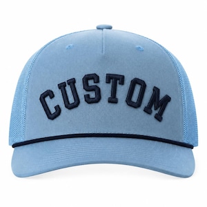 May include: A light blue trucker cap featuring a curved peak and a mesh back panel. The front panel displays the word CUSTOM in dark navy blue embroidered block lettering, accented by a thin dark navy blue cord trim across the base.