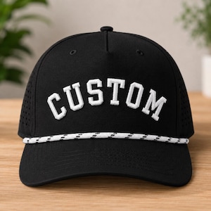 Puede incluir: Gorra de b&eacute;isbol negra con la palabra "CUSTOM" bordada en blanco. Tiene un detalle de cuerda blanca y negra y un panel lateral perforado. Un accesorio de moda.