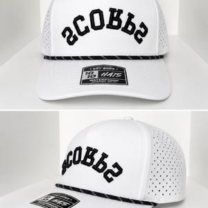 K&ouml;nnte beinhalten: Wei&szlig;e Baseballkappe mit schwarzem, gesticktem Text "SCOBBS". Die Kappe hat ein perforiertes Mesh-Seitenteil, ein schwarzes Seildetail und einen gebogenen Schirm. Ein Etikett mit Text ist am Schirm befestigt.