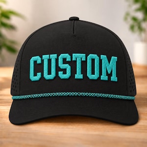 以下が含まれることがあります： 前面に太字のティール色のカレッジスタイルフォントで「CUSTOM」という文字が刺繍された黒い野球帽。キャップのベース部分にはお揃いのティール色の編み込みロープのディテールがあり、側面には通気孔のあるパネルが施されています。