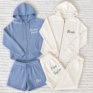 Puede incluir: Dos conjuntos de ropa de descanso a juego. Uno es un conjunto azul con sudadera con cremallera y pantalones cortos con el texto "Bridesmaid". El otro es un conjunto crema con sudadera con cremallera y pantalón de chándal con el texto "Bride" y "Mrs. Taylor".
